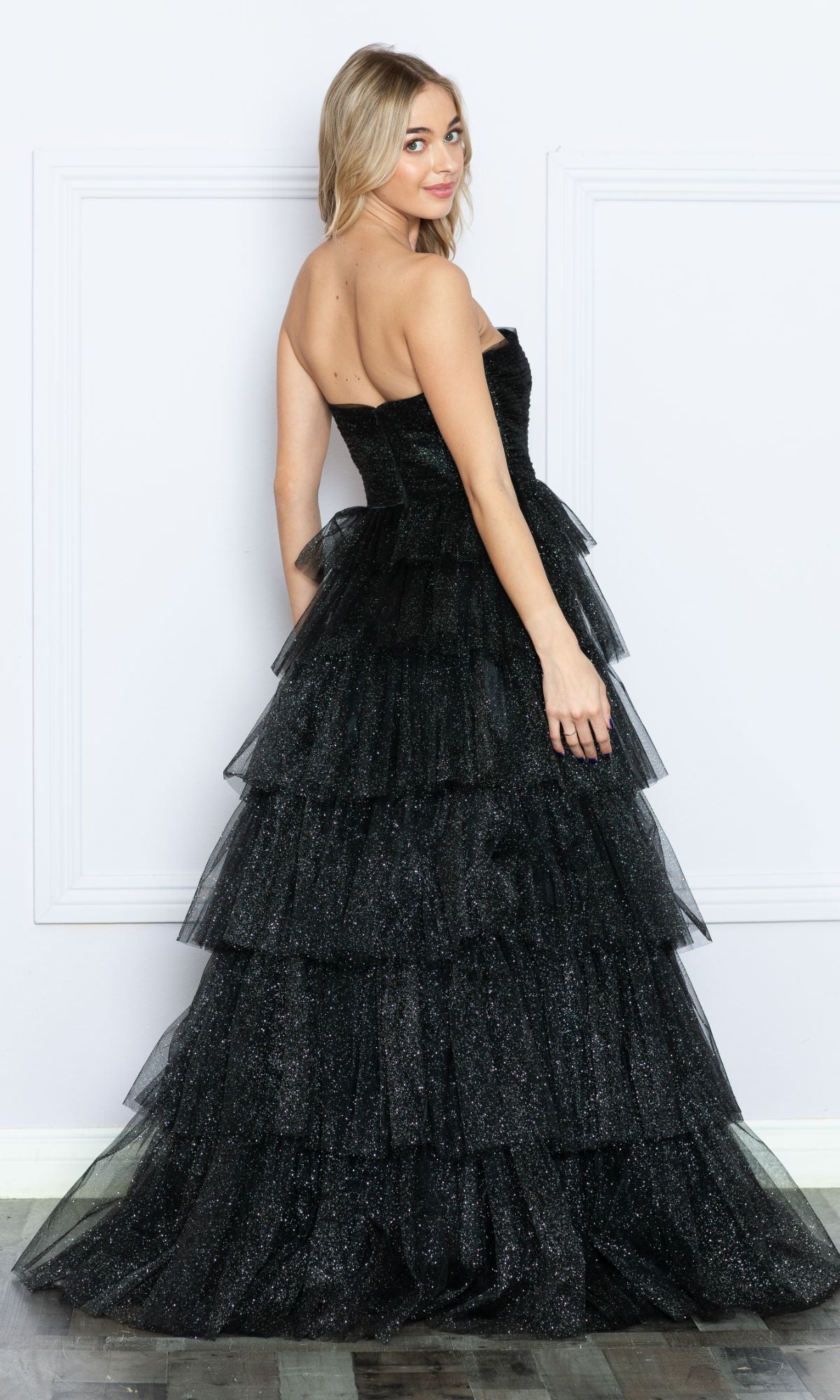A woman wears the Strapless Long Tiered Glitter Prom Ball Gown 9386, a black, floor-length, strapless evening dress with layered ruffle details and glitter tulle, posing with her back slightly turned.